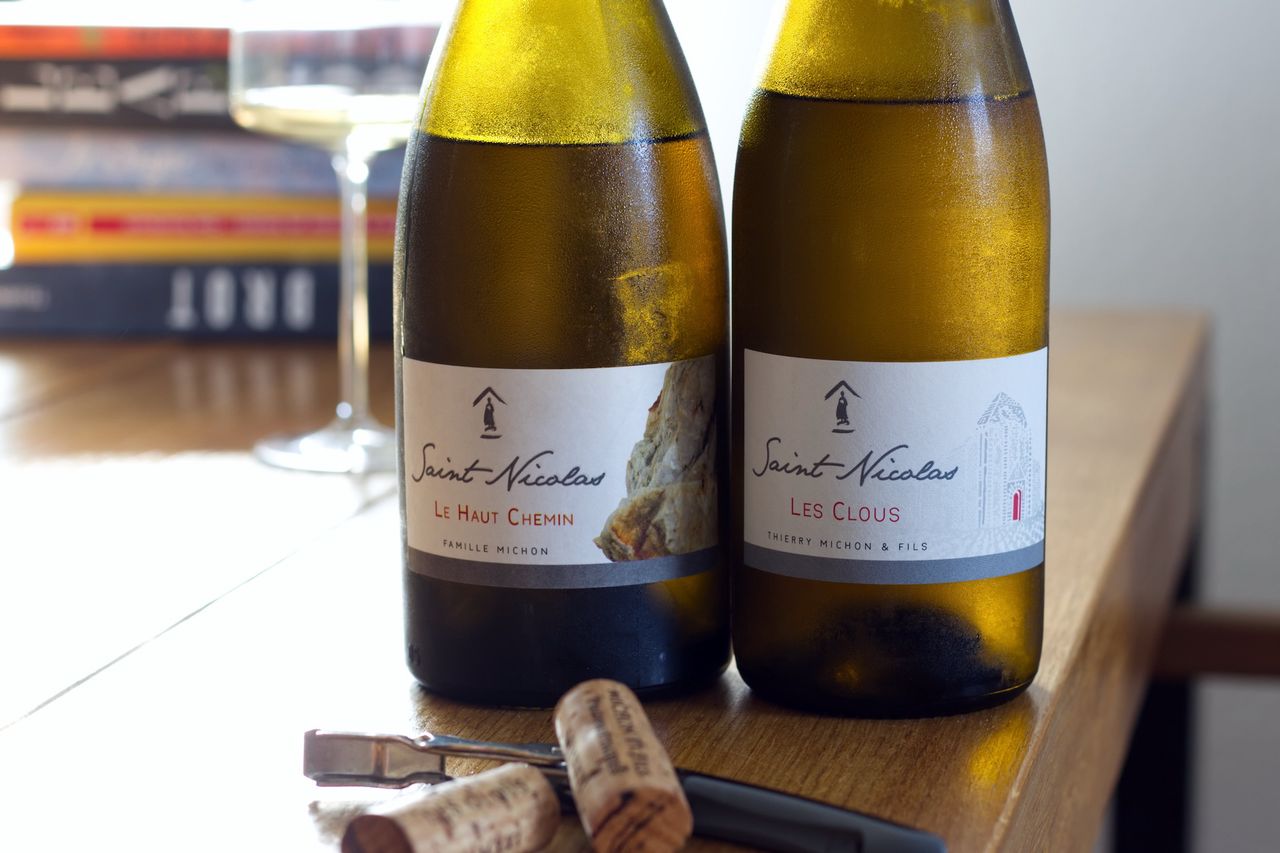 A bottle of Les Clous 2022 and a bottle of Le Haut Chemin 2021 of Domaine Saint Nicolas on a wooden table with a wine glass and books in the background, and corks with a corkscrew in the foreground.