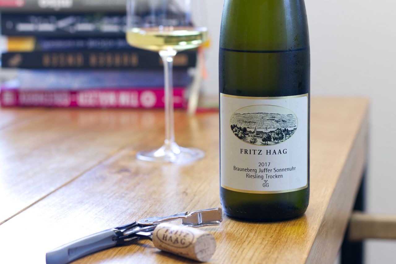 A bottle of Brauneberg Juffer Sonnenuhr Großes Gewächs 2017 from the Fritz Haag winery on a wooden table, with a wine glass and books in the background, and a cork and corkscrew in the foreground.