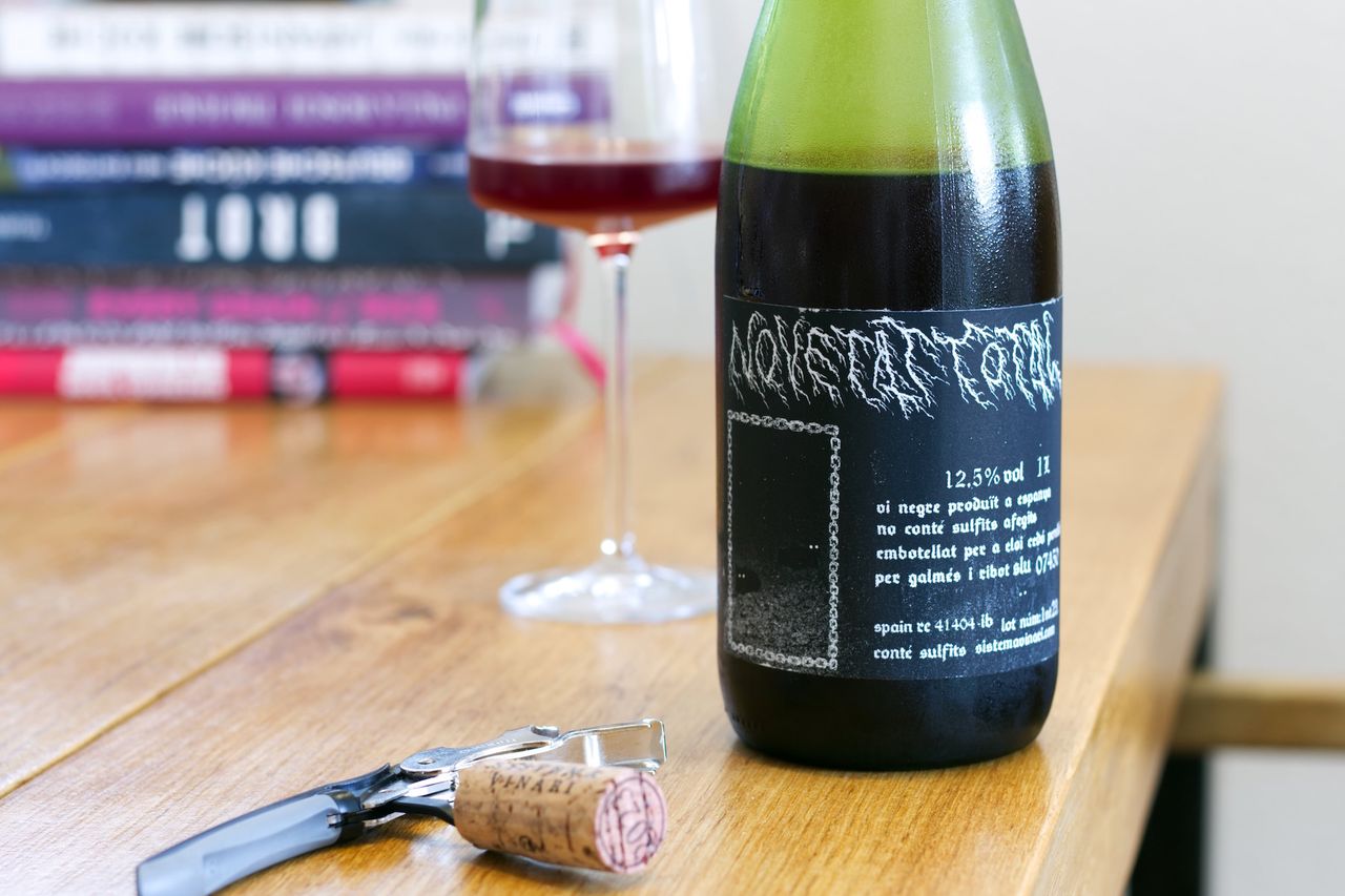 A bottle of Novetat Total 2023 from Sistema Vinari on a wooden table. The label resembles a black metal album cover. In the background, there is a wine glass and books, and in the foreground, there is a cork with a waiter's knife.