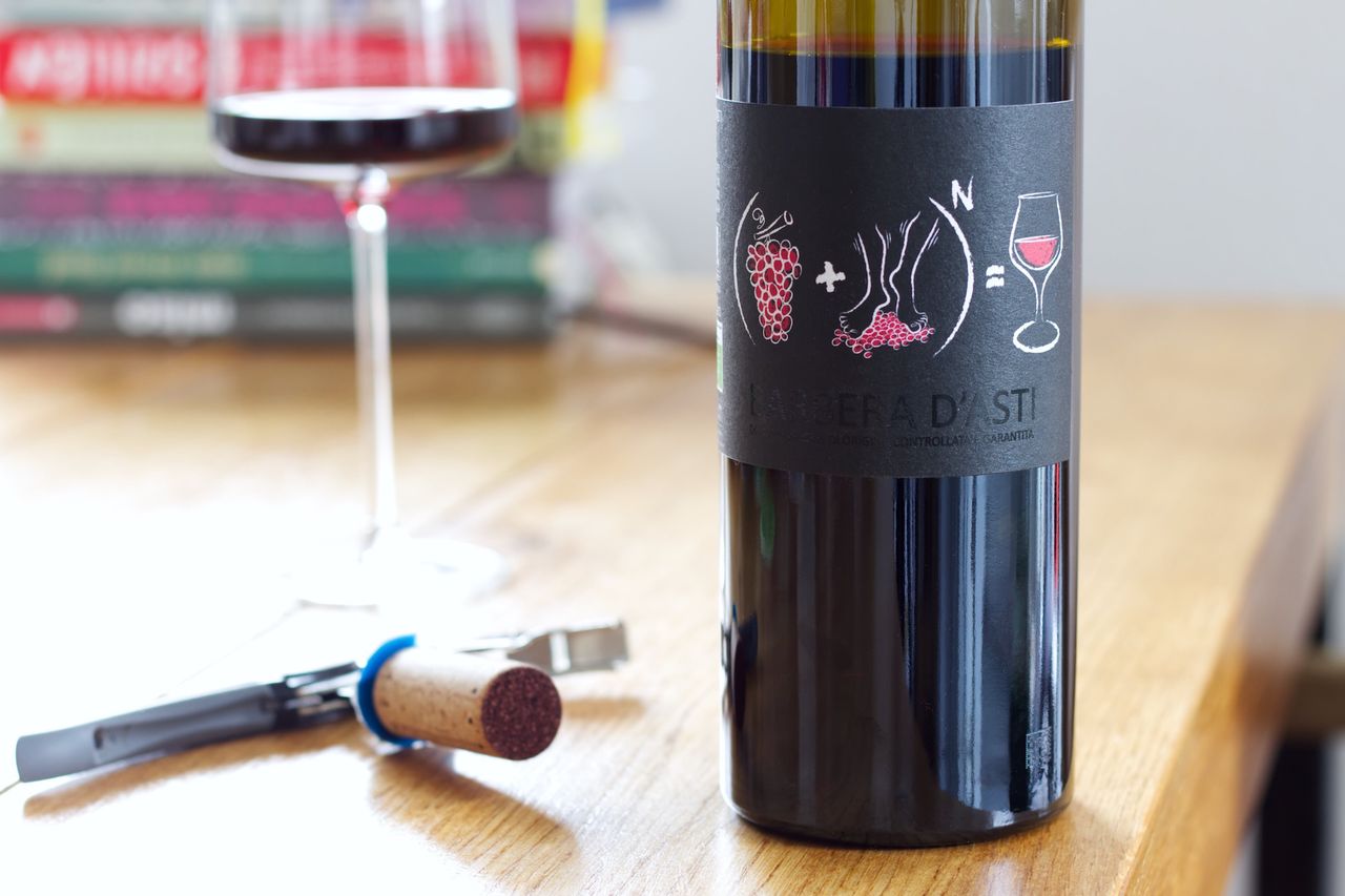 A bottle of Barbera d’Asti wine on a wooden table. The label on the bottle is black and features a formula with grapes and feet crushing the grapes, and on the other side of the equation, a wine glass. In the background, there is a filled wine glass and blurred cookbooks. In front of the bottle, a cork and a waiter’s corkscrew lie on the table.