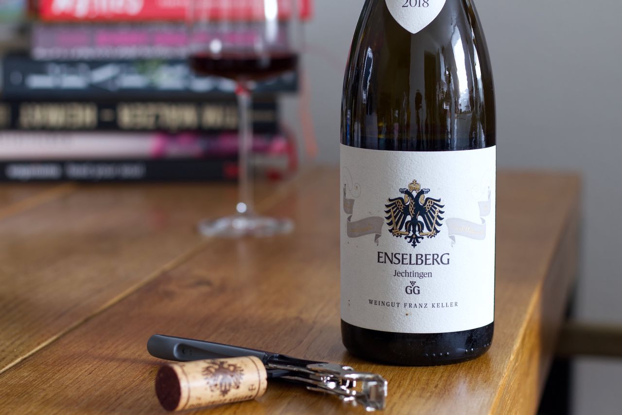 A bottle of Enselberg Spätburgunder from the 2018 vintage by Weingut Franz Keller on a wooden table. In the background, there is a wine glass and stacks of books. In front of the bottle, the cork lies on a waiter’s corkscrew.