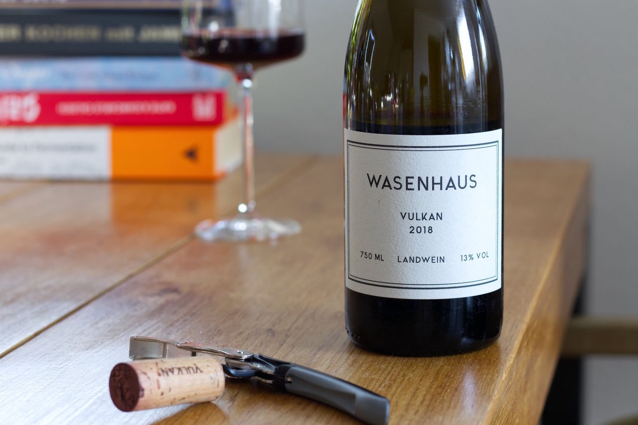 A bottle of wine from Weingut Wasenhaus on a wooden table. The minimalist label features only the wine name Vulkan, the vintage, the designation Landwein, and the winery's name in black text on a white background. In the background, there is a wine glass and stacks of books. In front of the bottle, there's the cork on a waiter's corkscrew.