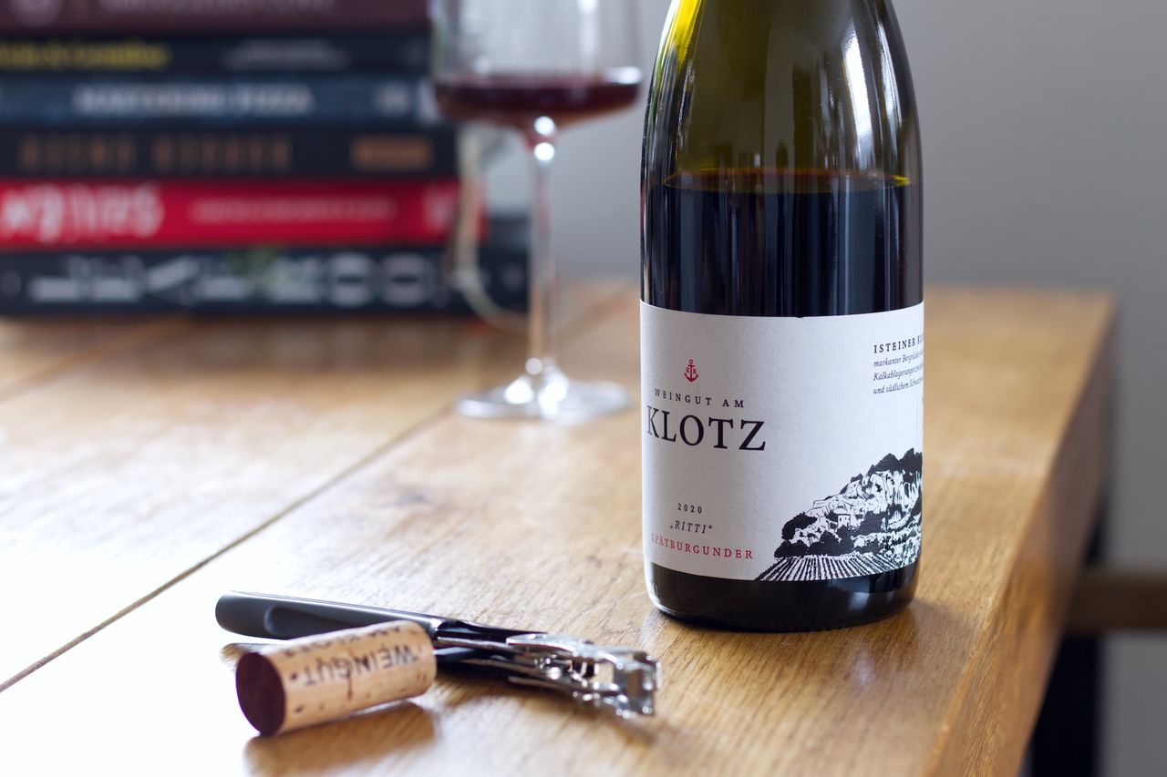 A bottle of wine from Weingut am Klotz on a wooden table. The label features the namesake Klotz, a mountain in southern Baden. In the background, there is a wine glass and a stack of books. In front of the bottle, the cork lies with a waiter's corkscrew.