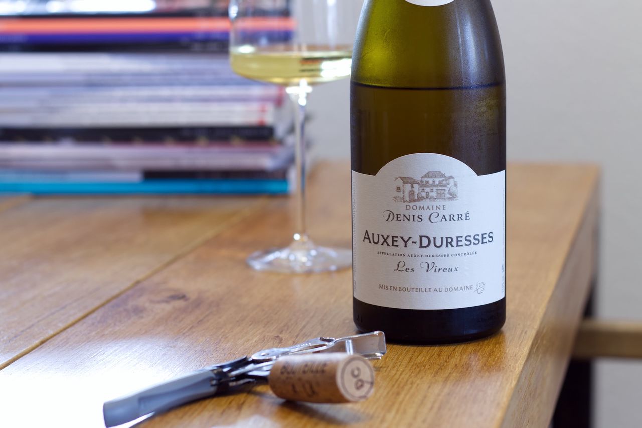 A bottle of Auxey-Duresses Les Vireux 2021 from Domaine Denis Carré on a wooden table. In the background, a wine glass stands in front of a stack of cookbooks, while in the foreground, a cork lies pinned to a sommelier knife.