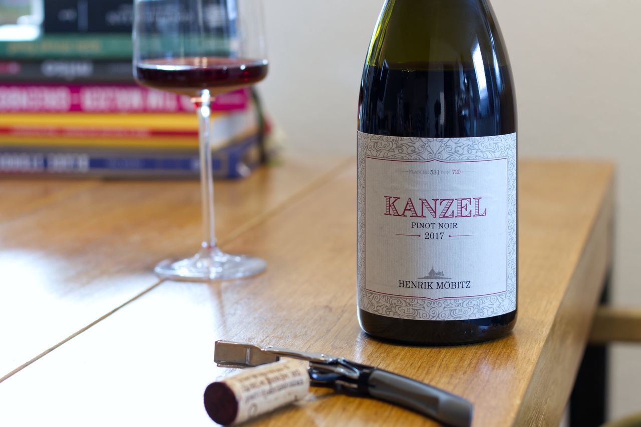 A bottle of wine from the Kanzel vineyard by Henrik Möbitz on a wooden table. In the background, a wine glass and stacks of books can be seen. In front of the bottle, the cork rests on a waiter's knife.