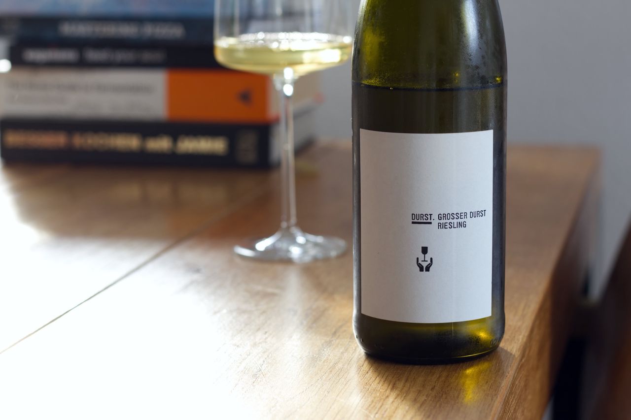 A bottle of Grosser Durst 2018 Riesling by Andreas Durst on a wooden table. In the background, a wine glass and stacks of books are visible.