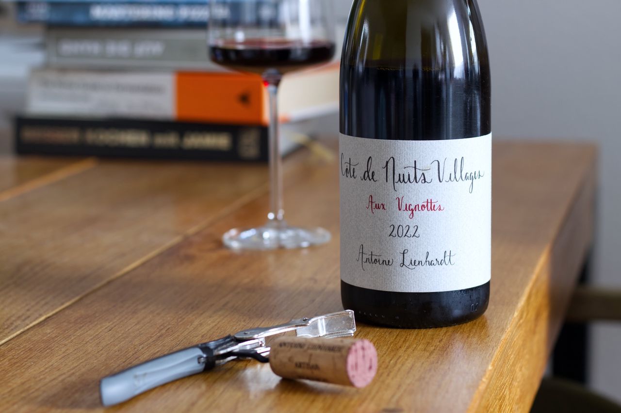 A bottle of wine from Antoine Lienhardt from the year 2022. The label features Côte de Nuits Villages and Aux Vignottes in cursive script. In the background, there's a wine glass and a stack of books. In front of the bottle, the cork rests on the waiter's knife.