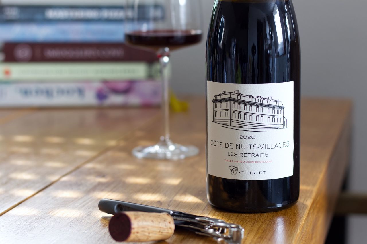 A bottle of Les Retraits Côte de Nuits Villages 2020 from Camille Thiriet on a wooden table. The label outlines a building. In the background, a wine glass and a stack of books can be seen. In front of the bottle, the cork rests on a waiter's knife.