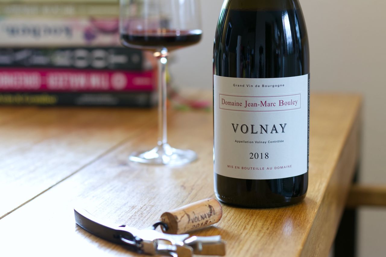 A bottle of Volnay 2018 from Domaine Jean-Marc Bouley on a wooden table. In the background, a filled wine glass stands in front of a stack of cookbooks, while in the foreground, a cork on a sommelier knife.