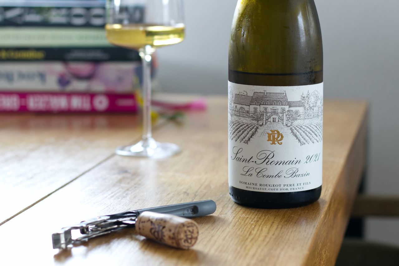 A bottle of Saint-Romain La Combe Bazin 2021 from Domaine Denis Rougeot on a wooden table. In the background, a filled wine glass stands in front of a stack of cookbooks, while in the foreground, the matching cork is stuck on a sommelier knife.