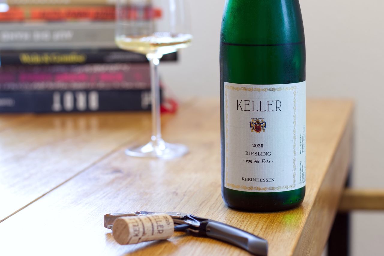 A bottle of Riesling Von der Fels from Weingut Keller on a wooden table. In the background, a wine glass and stacks of books are visible. In front of the bottle, the cork lies stuck to a waiter's knife.