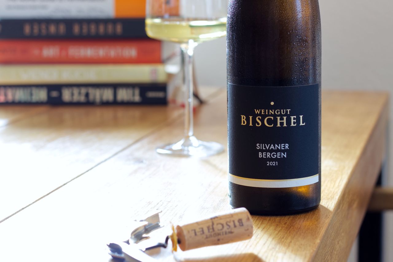 A bottle of Silvaner Bergen 2021 from Weingut Bischel on a wooden table. In the background, a filled wine glass stands in front of a stack of cookbooks, while in the foreground, the matching cork lies stuck to a waiter’s corkscrew.