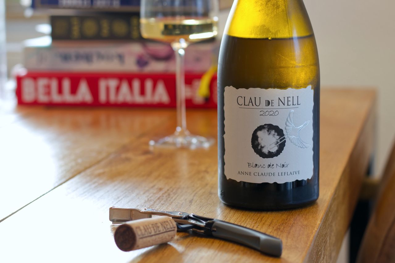 A bottle of Blanc de Noir 2020 from Clau de Nell on a wooden table. In the background, a wine glass and stacks of books can be seen. In front of the bottles, the cork of the bottle is attached to a waiter's corkscrew.