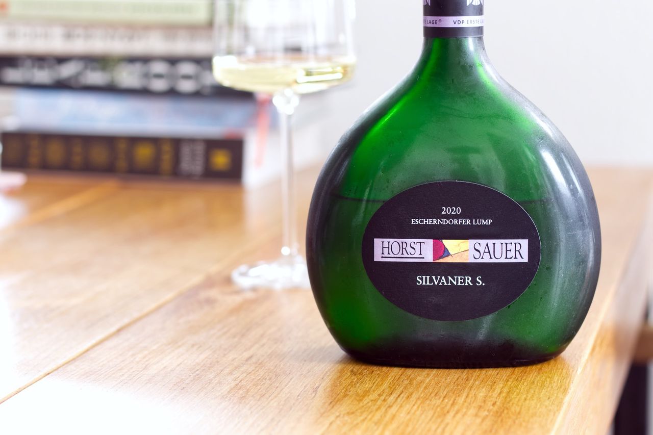 A Bocksbeutel bottle of Silvaner S wine from the Escherndorfer Lump vineyard by Horst Sauer on a wooden table. In the background, a wine glass and stacks of books can be seen.