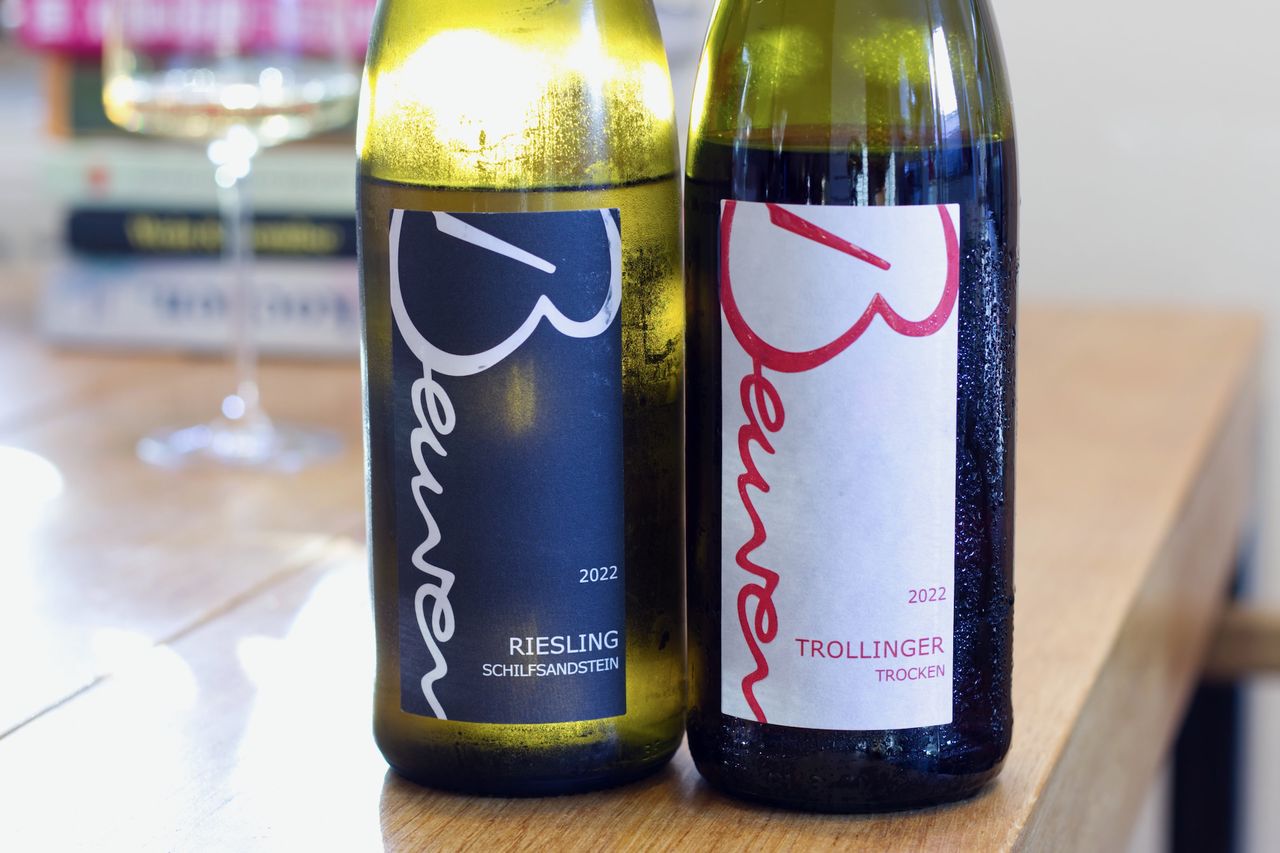 Two bottles of wine from Weingut Beurer stand on a wooden table, backlit. In the background, a wine glass and a stack of books can be seen.