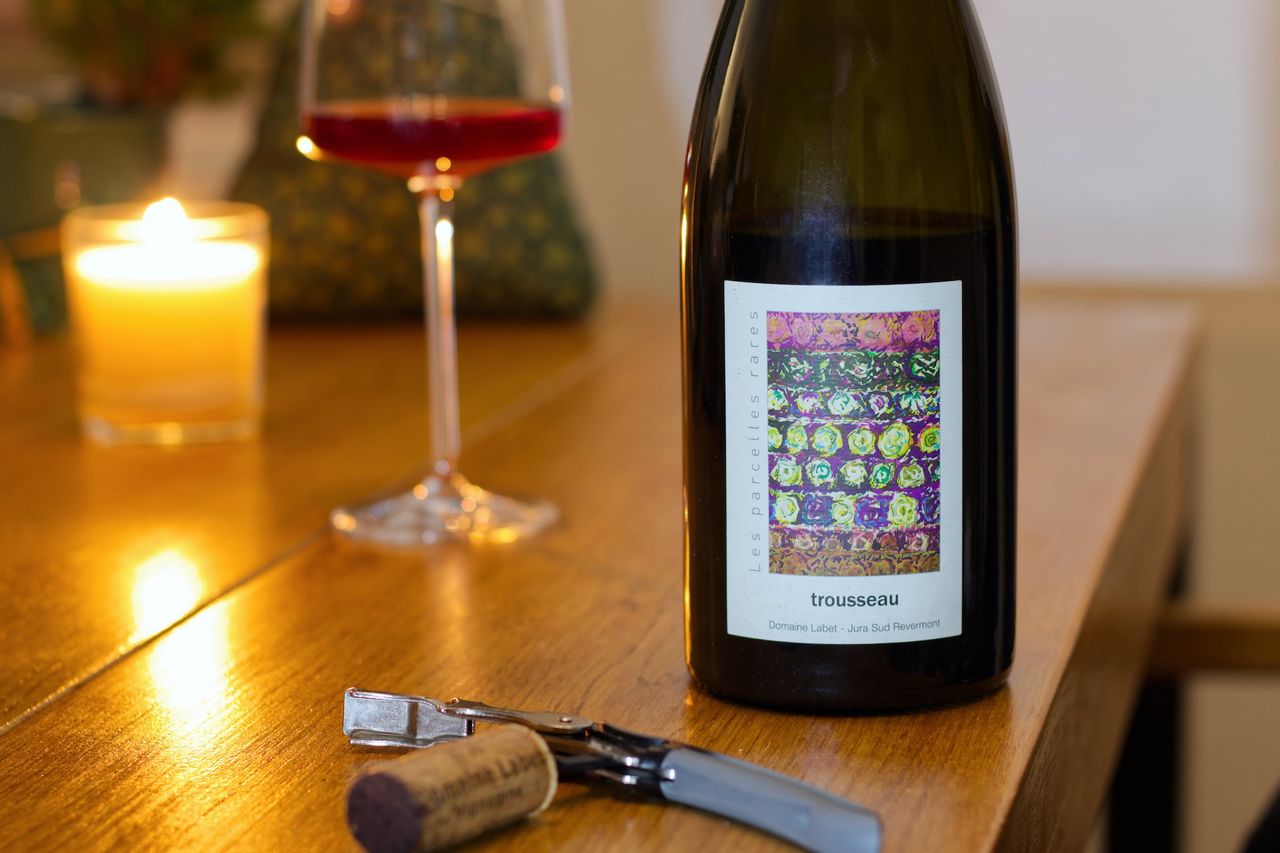 A bottle of Trousseau from Domaine Labet on a wooden table. In the background, there’s a fabric Christmas tree and a candle with a filled wine glass, while in the foreground, the matching cork is stuck to a waiter’s corkscrew.