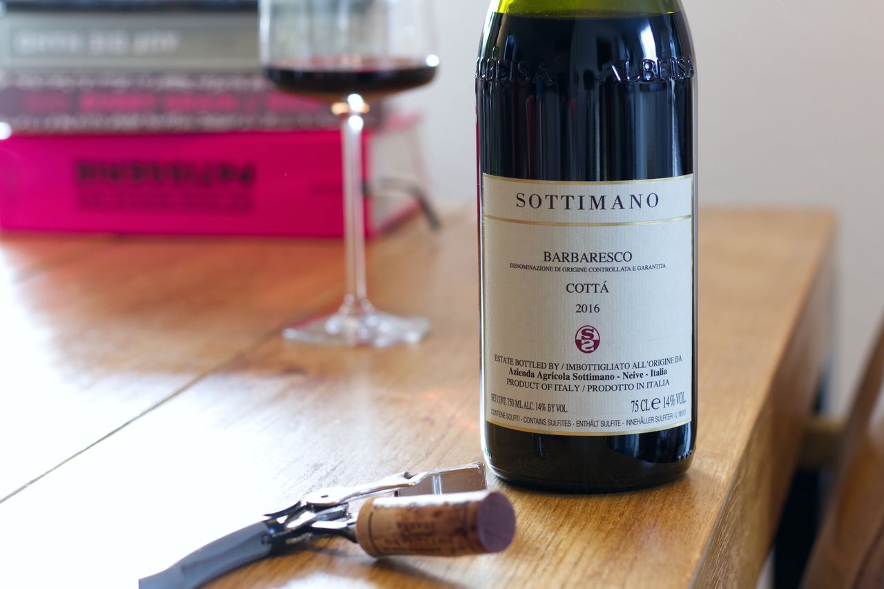 A bottle of wine from Sottimano stands on a wooden table. In the background, a wine glass and stacks of books are visible. In front of the bottle, the cork from the bottle lies next to the waiter’s corkscrew.