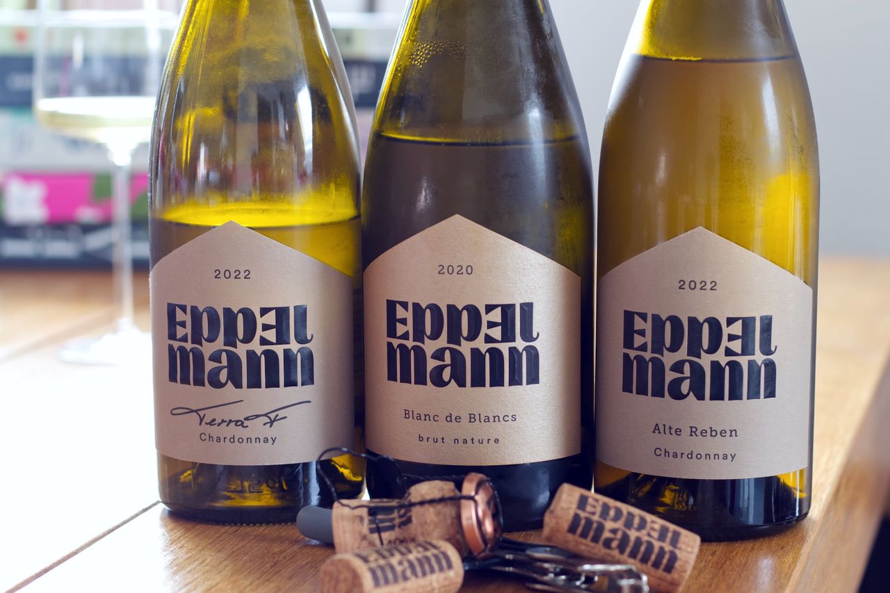 Three bottles of wine from Weingut Eppelmann on a wooden table. In the background, a filled wine glass stands in front of a stack of cookbooks, while in the foreground lie three corks and the waiter’s corkscrew.