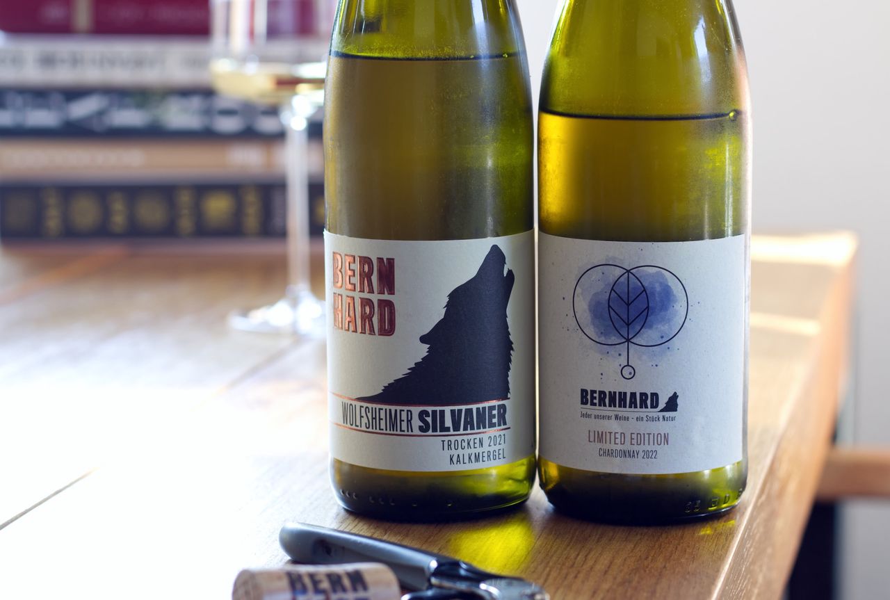 On a wooden table stand two bottles of wine from Weingut Bernhard. A Chardonnay in a limited edition and a Silvaner. In the background, a wine glass and a stack of books can be seen. In front of the bottles lie a cork and a waiter’s corkscrew.