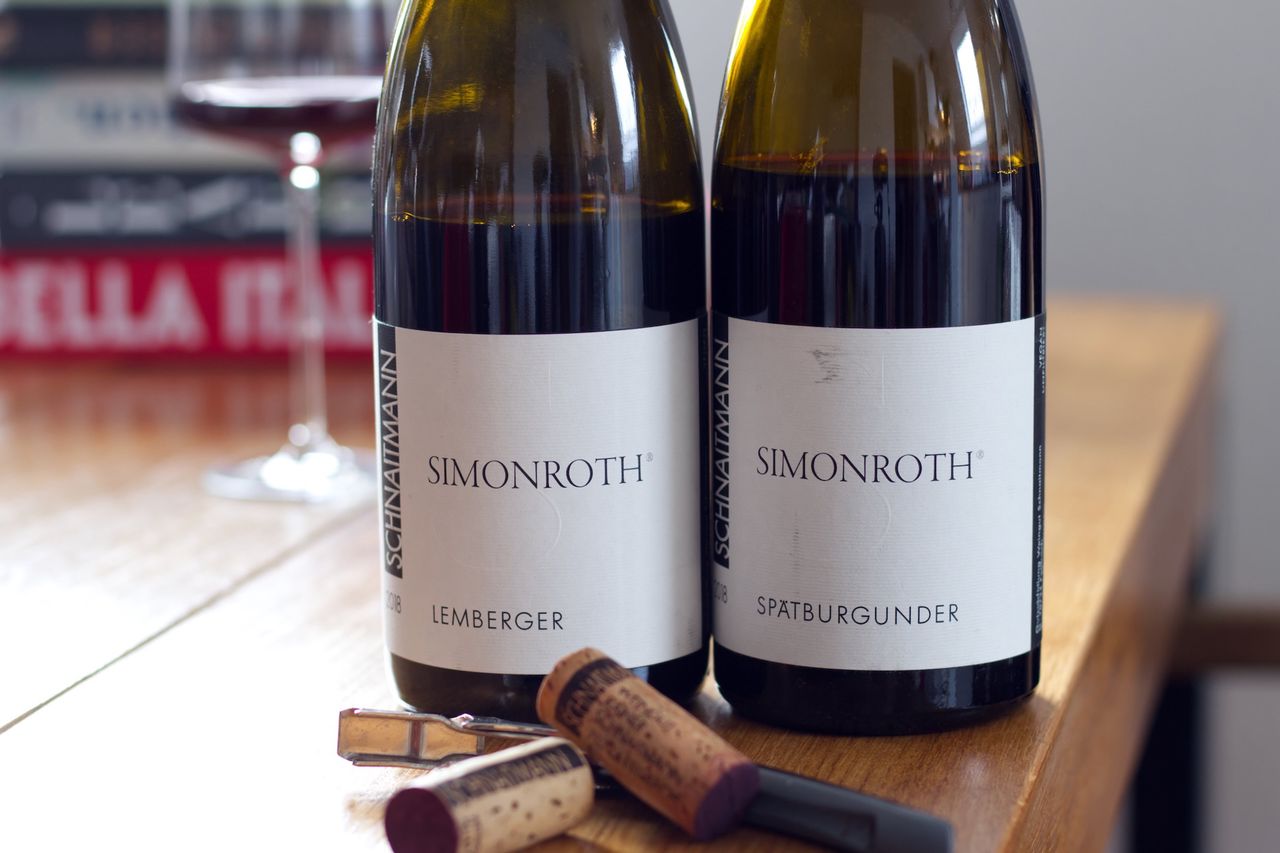 On a wooden table, there are two bottles of wine from Weingut Schnaitmann. One is a Spätburgunder and the other is a Lemberger, both from the Simonroth series. In the background, a wine glass and a stack of books are visible. In front of the bottles lie corks and a waiter’s corkscrew.