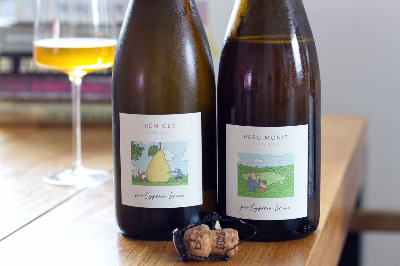 On a wooden table, there are two bottles of fruit sparkling wine from Cyprien Lireux. On one of the labels, someone is tapping a glass of Poiré from an oversized pear. In the background, a wine glass and a stack of books are visible.