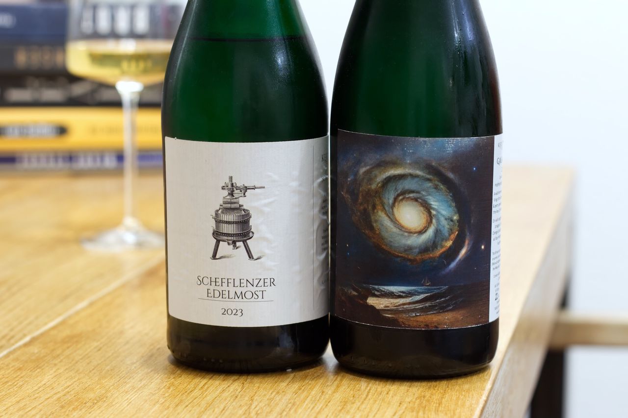 On a wooden table, there are two bottles of fruit sparkling wine from Kertelreiter. One label shows a traditional basket press, while the other displays a galaxy. In the background, a wine glass and a stack of books are visible.