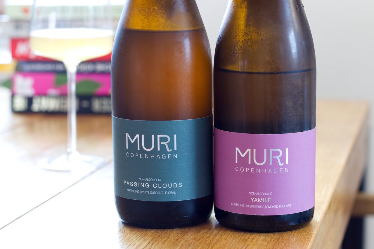 On a wooden table, there are two bottles of non-alcoholic sparkling drinks from Muri. One is Yamilé and the other is Passing Clouds. In the background, a wine glass and a stack of books are visible.