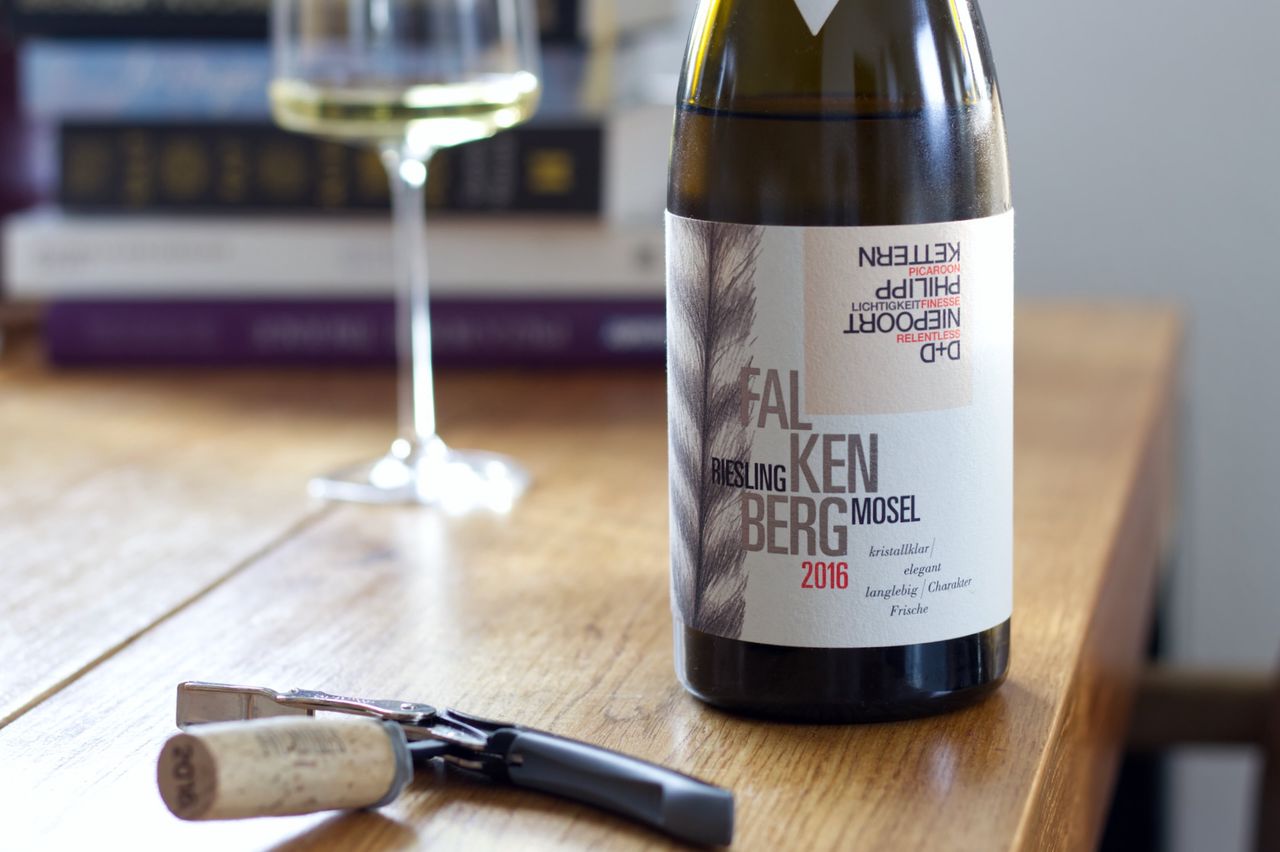 A bottle of Falkenberg 2016 from Fio Wines stands on a wooden table. In the background, a wine glass and a stack of books can be seen, in the foreground lies the cork on the waiter's knife.