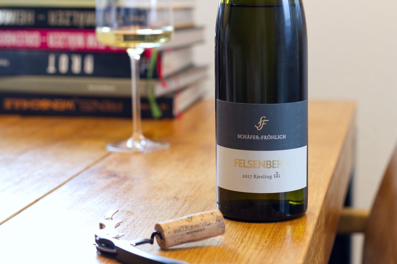 On a wooden table stands a bottle of Felsenberg 2017 from Schäfer-Fröhlich. In the background, a wine glass and a stack of books can be seen, while in the foreground, the cork lies on a sommelier's knife.