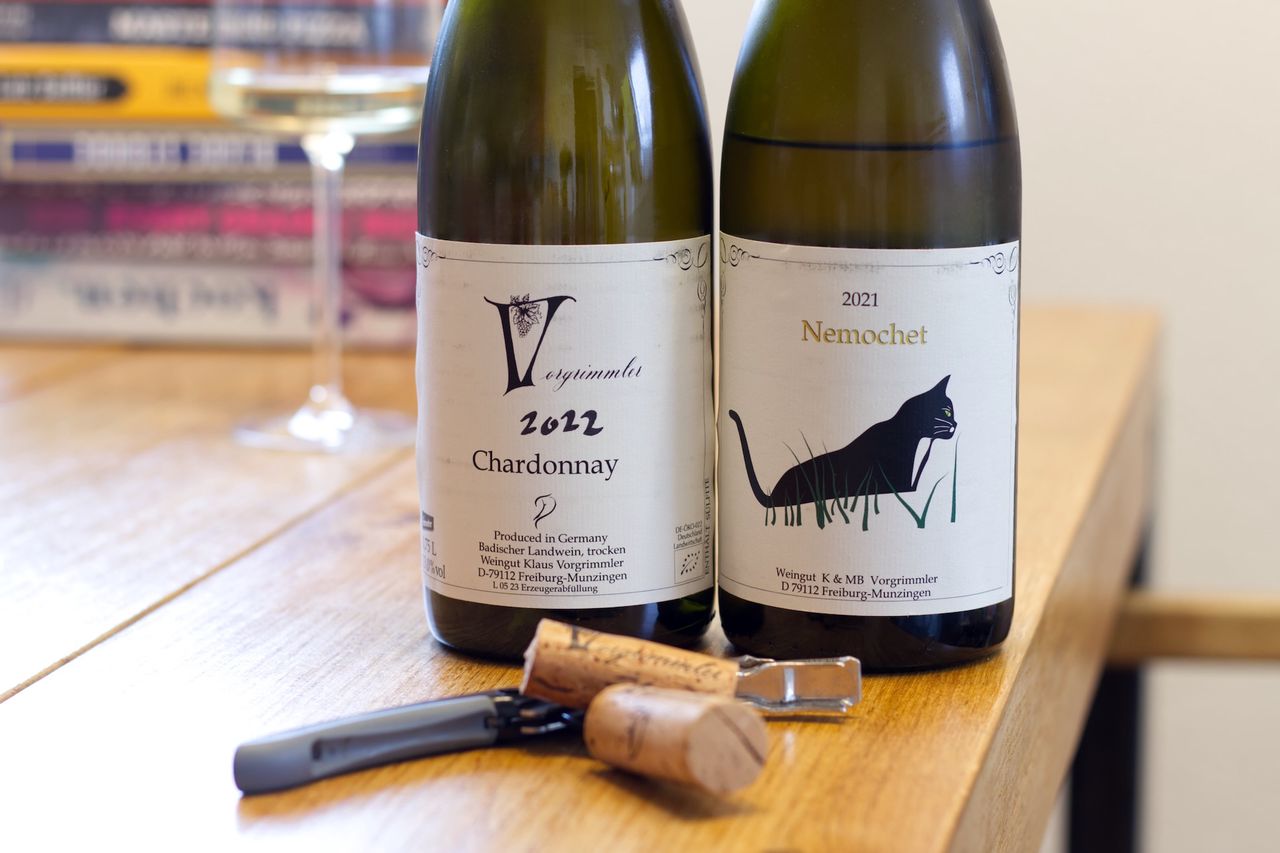 On a wooden table, there are two bottles of wine from Weingut Vorgrimmler. A Chardonnay 2022 and a Nemochet 2021 with a cat on the label. In the background, a wine glass and a stack of books are visible.