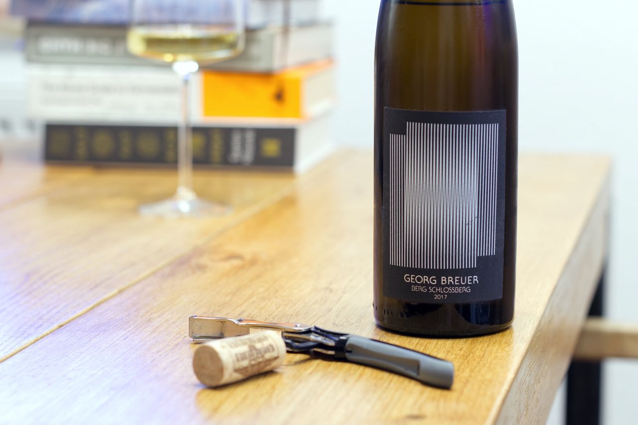 On a wooden table stands a bottle of Berg Schlossberg 2017 from Georg Breuer with an artist label. In the background, a wine glass and a stack of books can be seen, while in the foreground, the cork lies on a sommelier's knife.